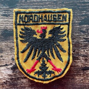 May include: Embroidered patch with the word "NORDHAUSEN" at the top. The patch features a black eagle with red accents on a yellow background, framed by a black border. The patch is likely a collectible or souvenir.