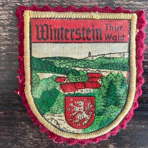 Op de afbeelding: Een schildvormige stoffen embleem met een landschapsillustratie van Winterstein Thür. Wald met gebouwen met rode daken, groene bomen en een klein rood wapenschild met een witte leeuw. Het embleem heeft een gouden geborduurde rand en rode schulprand.