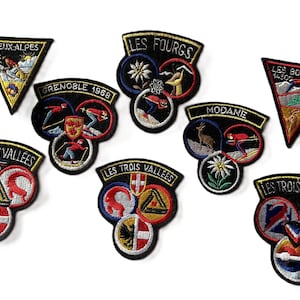 Peut inclure: Un ensemble d'écussons brodés représentant des logos et noms de stations de ski. Les écussons, de formes variées (triangles, cercles), affichent des textes tels que "DEUX ALPES", "LES FOURGS" et "LES TROIS VALLEES".