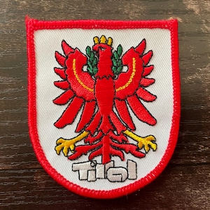 Könnte beinhalten: Gestickter Aufnäher mit einem roten Adler-Emblem, gelben Akzenten und grünen Blättern auf weißem Hintergrund. Der Aufnäher hat einen roten Rand und das Wort "Tirol" in weißen Blockbuchstaben.