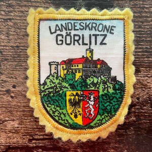 May include: A rectangular fabric patch with scalloped yellow edges featuring the text LANDESKRONE GÖRLITZ in black. The design depicts a building with a red roof atop a green hill, with a shield showing a black eagle and a red lion below.