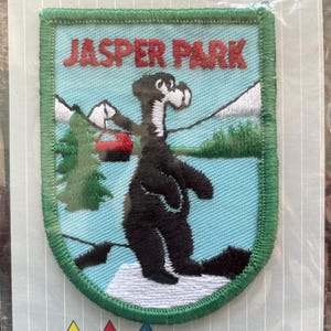 May include: A rectangular embroidered patch with a green border featuring the text JASPER PARK in red. The design depicts a black bear standing on a white snowy surface against a light blue background with green pine trees and a red cable car.