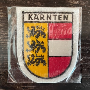 May include: A rectangular patch with the word "KÄRNTEN" at the top. The patch is divided into three sections: a yellow field with black lions, a white field, and a red field. The patch is in a clear plastic wrapper.