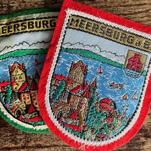 Peut inclure: Deux écussons brodés portant le texte MEERSBURG et MEERSBURG A.B. Ils représentent une scène de ville au bord d'un lac avec un château, des voiliers sur l'eau bleue et des montagnes en arrière-plan, bordés de vert et de rouge.