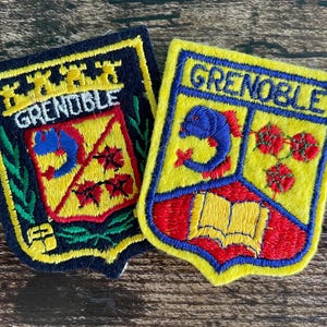 Può includere: Due toppe ricamate a forma di scudo con la scritta GRENOBLE. La toppa a sinistra ha un bordo blu scuro con uno stemma giallo, rosso e blu. La toppa a destra ha un bordo giallo con uno stemma blu, rosso e giallo che raffigura un pesce e un libro.