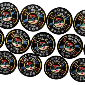 May include: A collection of circular black embroidered patches featuring a skier in a red jacket and blue pants, yellow crossed skis, and white snowflake borders. Each patch displays a unique French ski resort name and elevation text.