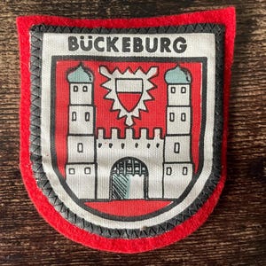 Peut inclure: Un écusson en tissu en forme de bouclier avec une bordure en feutre rouge. Le centre présente un blason blanc avec le texte BÜCKEBURG en haut. Le motif montre un fond rouge avec deux tours blanches, une porte centrale et un petit écu rouge.