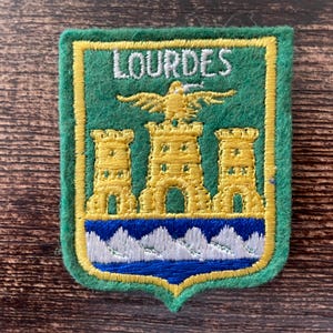 May include: Embroidered patch with a green background and a yellow border. The patch features the word "LOURDES" in white, above a gold emblem of a bird and three gold castle towers. A blue and white wave design is at the bottom.