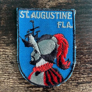 May include: A shield-shaped embroidered patch with a blue background featuring the text ST. AUGUSTINE FLA. in white. The design includes a black silhouette of a sailing ship and a profile of a figure wearing a helmet with a red plume and collar.