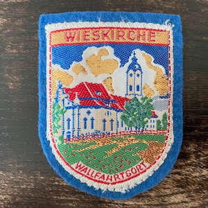 Puede incluir: Parche azul en forma de escudo con el texto "WIESKIRCHE" en la parte superior. El parche presenta una imagen bordada de una iglesia con tejado rojo y alto campanario. El texto "WALLFAHRTSORT" está en la parte inferior.