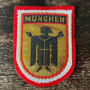 May include: A rectangular patch with a red border, white and gold trim, and a gold background. The patch features a black emblem and the word "MUNCHEN" in gold letters. The patch is likely a souvenir or emblem.