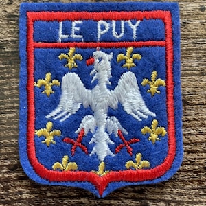 May include: A blue felt patch shaped like a shield with a red embroidered border. It features the white text LE PUY at the top, a central white eagle with red talons, and eight small yellow fleur-de-lis symbols arranged around the eagle.