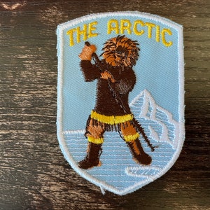 Könnte beinhalten: Gestickter Aufnäher mit hellblauem Hintergrund und weißem Rand. Der Aufnäher zeigt den Text "THE ARCTIC" in Gelb. Eine braune Figur mit Pelzmütze und gelben Akzenten hält einen Speer. Ein weißer Berg ist im Hintergrund.