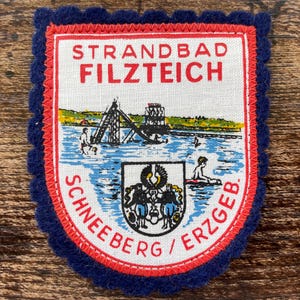 Puede incluir: Un parche de tela rectangular con un borde de fieltro azul oscuro festoneado. El centro blanco presenta un diseño con borde rojo con el texto STRANDBAD FILZTEICH y SCHNEEBERG/ERZGEB. Representa una escena de lago con un tobogán y un escudo.