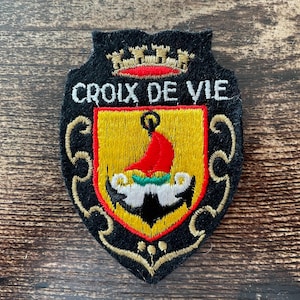 Peut inclure: Un écusson brodé noir en forme de blason portant l'inscription blanche CROIX DE VIE. Le centre présente un écu jaune avec une voile rouge, des détails verts et des vagues blanches. Une couronne dorée surmonte le texte avec des volutes dorées.