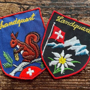 May include: Two shield-shaped embroidered patches with red borders. The blue patch features a squirrel on a branch with a pinecone and a Swiss flag. The black patch displays a Swiss flag, mountains, and an edelweiss flower. Both read Landquart.