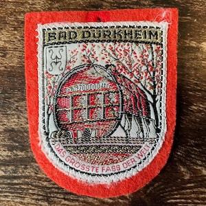 Puede incluir: Un parche rectangular de fieltro rojo con un diseño tejido de un gran barril de vino. El texto dice BAD DURKHEIM en la parte superior y DAS GROSSTE FASS DER WELT en la parte inferior. El gráfico central muestra un barril con 1700000 LTR.