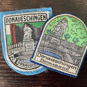 Op de afbeelding: Twee geborduurde stoffen patches met het Donaueschingen Donauquelle monument. De linker patch heeft een lichtblauwe rand en de tekst DONAUESCHINGEN. De rechter patch heeft een donkerblauwe rand en de tekst Donaueschingen Donauquelle.