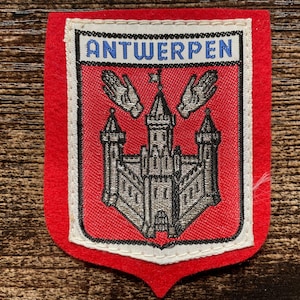 Op de afbeelding: Een rode vilten patch met een geborduurd schildontwerp. Het schild toont een grijs kasteel met twee handen erboven en de tekst ANTWERPEN in blauwe letters op een witte achtergrond aan de bovenzijde. De patch heeft een witte rand.