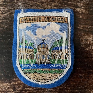 May include: A blue and white embroidered patch featuring a detailed image of a building with fountains and the text "BAYREUTH-EREMITAGE". The patch has a blue felt border and white stitching.