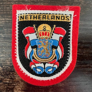 Può includere: Una toppa in feltro rosso con un emblema ricamato raffigurante una corona dorata, uno scudo blu con un leone d'oro e accenti rossi, bianchi e blu. La parola NETHERLANDS è ricamata in filo dorato sullo sfondo nero.