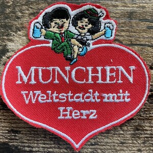 May include: A heart-shaped embroidered patch with a red background and white border. The top features two cartoon figures holding blue beer mugs. The text reads MUNCHEN Weltstadt mit Herz in white thread, centered on the red heart surface.