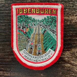 May include: Embroidered patch with a red border and white stitching. The patch features the text "IBBENBUREN" in gold lettering at the top. The center depicts a scenic view of a roller coaster and the text "SOMMERRODELBAHN MARCHENWALD".