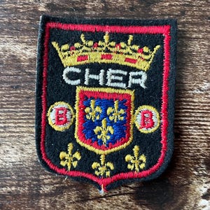 May include: A black embroidered patch in the shape of a shield with a red border. It features a gold crown above the white text CHER, a central blue shield with gold fleur-de-lis, two small circles with red B letters, and three gold fleur-de-lis.