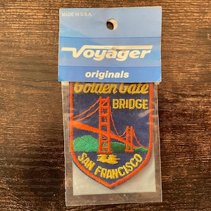 May include: A patch featuring the Golden Gate Bridge in San Francisco, California. The patch is in a clear plastic sleeve and has a blue background with an orange bridge design and green hills. The text "Voyager originals" is at the top.