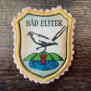 May include: A shield-shaped patch with the text "BAD ELSTER" above a black and white bird illustration. The bird stands on a green hill with a red and blue water feature. The patch has a gold border and a pink scalloped edge.
