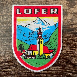 Könnte beinhalten: Ein schildförmiger Aufnäher mit dem Schriftzug LOFER in schwarzen Buchstaben auf rotem Grund. Die zentrale Abbildung zeigt eine gelbe Kirche mit rotem Dach vor einer Landschaft aus grünen Hügeln und blau schneebedeckten Bergen.