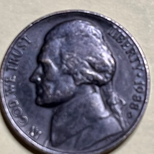 May include: A silver US nickel coin with the profile of Thomas Jefferson on the obverse side. The coin has the words "Liberty" and "In God We Trust" inscribed around the edge.