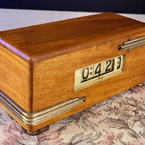 May include: A vintage wooden flip clock with a rectangular shape. The clock displays the time as 0:42. The clock has a light brown wooden exterior with silver-colored metal accents and small feet.