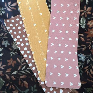 Puede incluir: Cuatro marcap&aacute;ginas rectangulares con varios dise&ntilde;os. Uno es marr&oacute;n con lunares blancos, otro es amarillo con la palabra "love" repetida, un tercero es rosa con flechas blancas y el &uacute;ltimo es marr&oacute;n con lunares blancos.