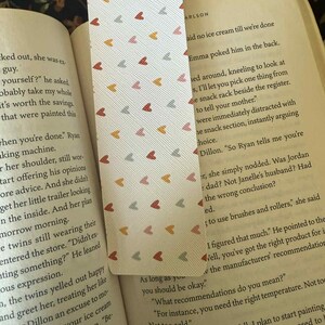 Puede incluir: Un marcap&aacute;ginas rectangular con fondo blanco y un patr&oacute;n repetido de peque&ntilde;os corazones en tonos rojos, naranjas y grises. El marcap&aacute;ginas est&aacute; dentro de un libro abierto.