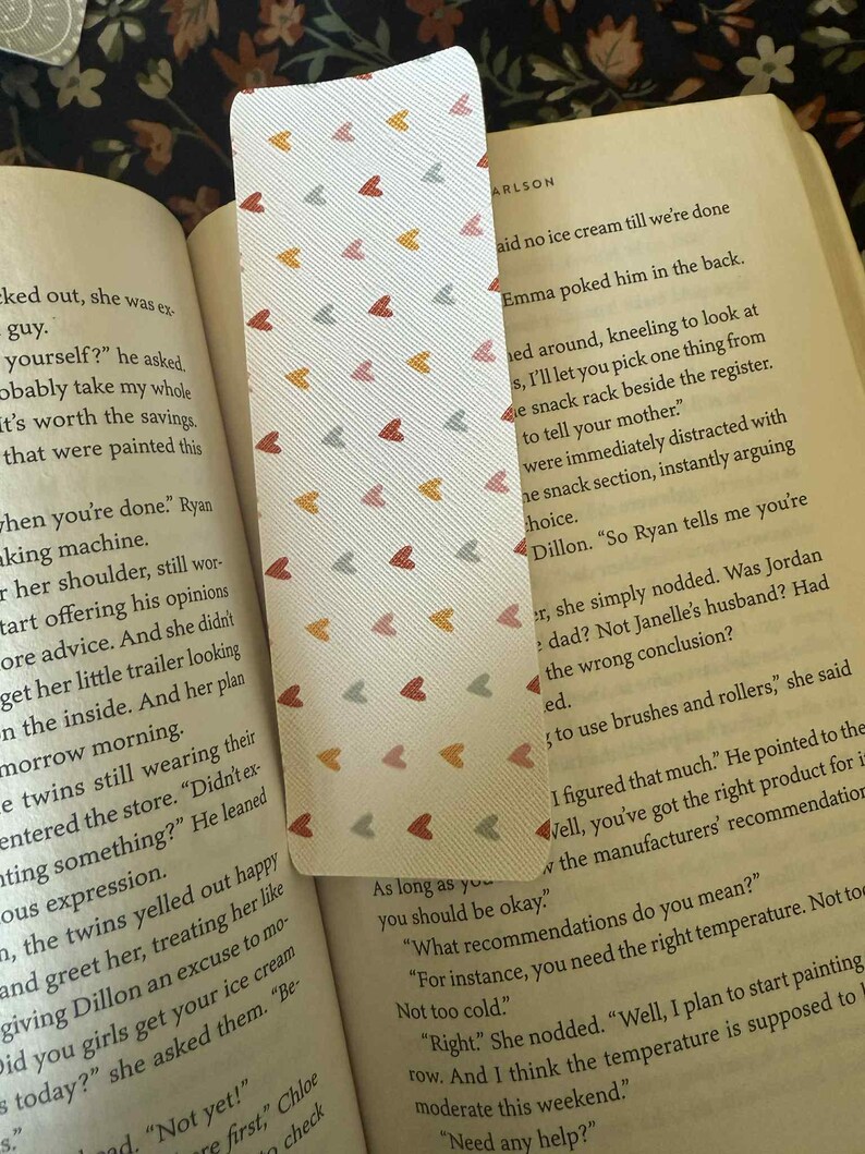 Puede incluir: Un marcap&aacute;ginas rectangular con fondo blanco y un patr&oacute;n repetido de peque&ntilde;os corazones en tonos rojos, naranjas y grises. El marcap&aacute;ginas est&aacute; dentro de un libro abierto.