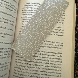 Puede incluir: Un marcap&aacute;ginas con estampado gris y blanco se coloca en un libro abierto. El marcap&aacute;ginas tiene una parte superior curvada y un dise&ntilde;o de c&iacute;rculos y corazones. Las p&aacute;ginas del libro son visibles, con texto y el n&uacute;mero 107.