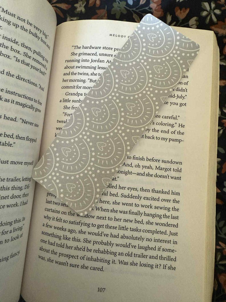 Puede incluir: Un marcap&aacute;ginas con estampado gris y blanco se coloca en un libro abierto. El marcap&aacute;ginas tiene una parte superior curvada y un dise&ntilde;o de c&iacute;rculos y corazones. Las p&aacute;ginas del libro son visibles, con texto y el n&uacute;mero 107.