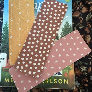 Puede incluir: Cuatro marcap&aacute;ginas rectangulares en tonos marr&oacute;n, rosa y amarillo, con patrones de puntos y corazones blancos. Los marcap&aacute;ginas est&aacute;n colocados encima de un libro con el t&iacute;tulo parcialmente visible. El fondo es un estampado floral.