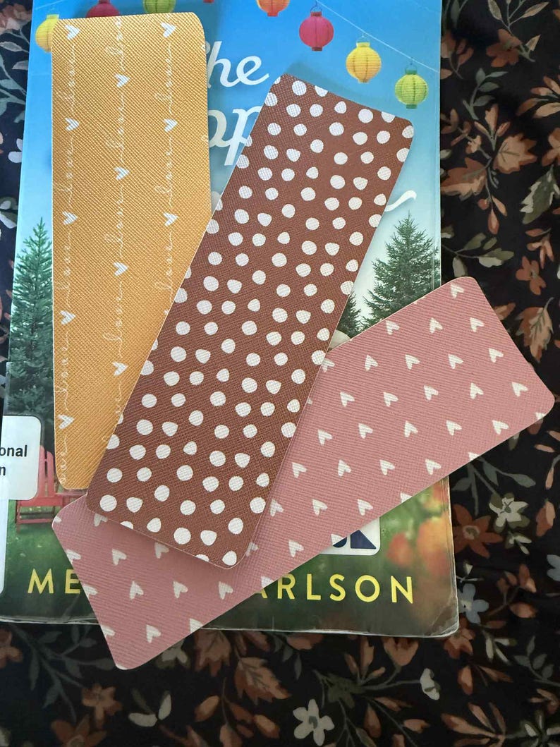 Puede incluir: Cuatro marcap&aacute;ginas rectangulares en tonos marr&oacute;n, rosa y amarillo, con patrones de puntos y corazones blancos. Los marcap&aacute;ginas est&aacute;n colocados encima de un libro con el t&iacute;tulo parcialmente visible. El fondo es un estampado floral.