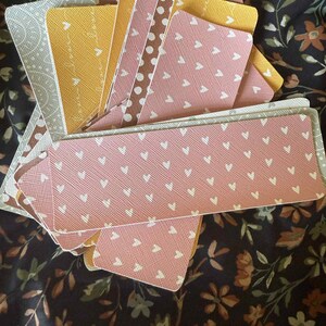 Puede incluir: Un abanico de marcap&aacute;ginas decorativos con varios dise&ntilde;os. Los marcap&aacute;ginas presentan dise&ntilde;os como corazones y lunares en tonos rosa, amarillo y marr&oacute;n. Los marcap&aacute;ginas est&aacute;n colocados sobre un fondo con estampado floral.