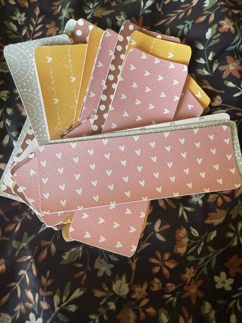 Puede incluir: Un abanico de marcap&aacute;ginas decorativos con varios dise&ntilde;os. Los marcap&aacute;ginas presentan dise&ntilde;os como corazones y lunares en tonos rosa, amarillo y marr&oacute;n. Los marcap&aacute;ginas est&aacute;n colocados sobre un fondo con estampado floral.