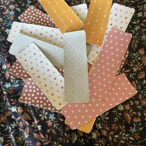 Puede incluir: Una colecci&oacute;n de marcap&aacute;ginas rectangulares con varios dise&ntilde;os, incluyendo lunares, corazones y dise&ntilde;os de cachemira. La paleta de colores incluye tonos de marr&oacute;n, rosa, amarillo y gris. Los marcap&aacute;ginas est&aacute;n colocados sobre una tela con estampado floral.