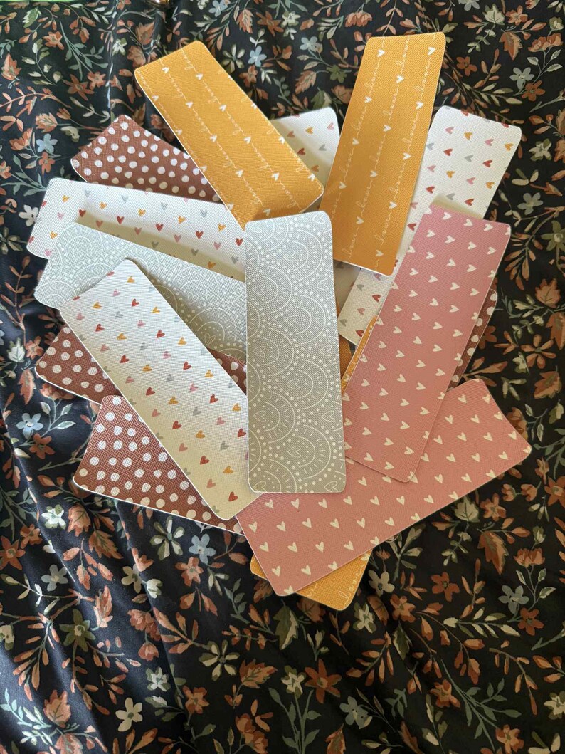 Puede incluir: Una colecci&oacute;n de marcap&aacute;ginas rectangulares con varios dise&ntilde;os, incluyendo lunares, corazones y dise&ntilde;os de cachemira. La paleta de colores incluye tonos de marr&oacute;n, rosa, amarillo y gris. Los marcap&aacute;ginas est&aacute;n colocados sobre una tela con estampado floral.