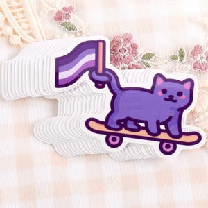 Puede incluir: Una pila de pegatinas troqueladas que muestran un gato morado sobre un monopatín color canela. El gato sostiene con la cola una bandera con franjas horizontales moradas, blancas y violetas. El diseño tiene un contorno morado oscuro.