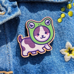 May include: A wooden pin featuring a cartoon dog wearing a green frog hat. The dog is white with purple spots and a purple tail. The pin is set against a blue denim background.