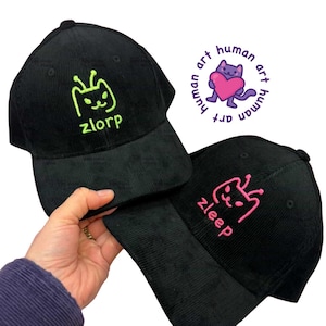 Puede incluir: Dos gorras de béisbol de pana negras. Una gorra tiene bordado verde neón con la palabra "zlorp" y un diseño de gato. La otra gorra tiene bordado rosa con la palabra "zleep" y un diseño similar. También es visible un logotipo morado y rosa.