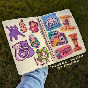 May include: Open notebook with colorful stickers. Stickers include a purple cat, a banana, a french fry, a can of beans, a cat with a frog hat, and text that says "I want power", "Yeah, I'm a GAMER", and "certified yapper".