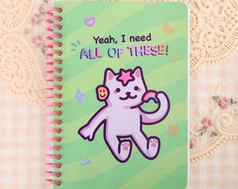 Puede incluir: Un cuaderno de espiral con una cubierta de rayas onduladas de color verde claro. El texto "Yeah, I need ALL OF THESE!" aparece sobre una ilustración de un gato morado claro con una estrella rosa en la cabeza, sosteniendo un arcoíris y un smiley.