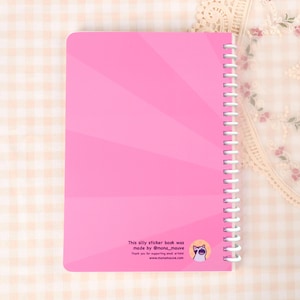May include: A pink spiral-bound notebook with a radiating design on the cover. The text "This silly sticker book was made by @mona_mauve" is printed at the bottom, along with a cat illustration and website address.